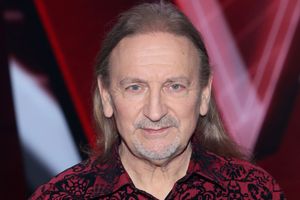 Mężczyzna z długimi włosami w kwiecistej koszuli, uśmiechający się do kamery.