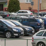 Parking pełen samochodów osobowych, z budynkiem w tle. Zielony znak parkowania w rogu.