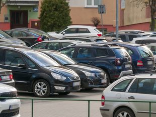 Parking pełen samochodów osobowych, z budynkiem w tle. Zielony znak parkowania w rogu.