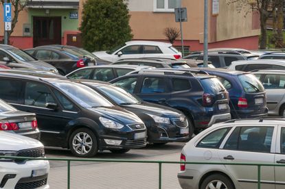 Parking pełen samochodów osobowych, z budynkiem w tle. Zielony znak parkowania w rogu.