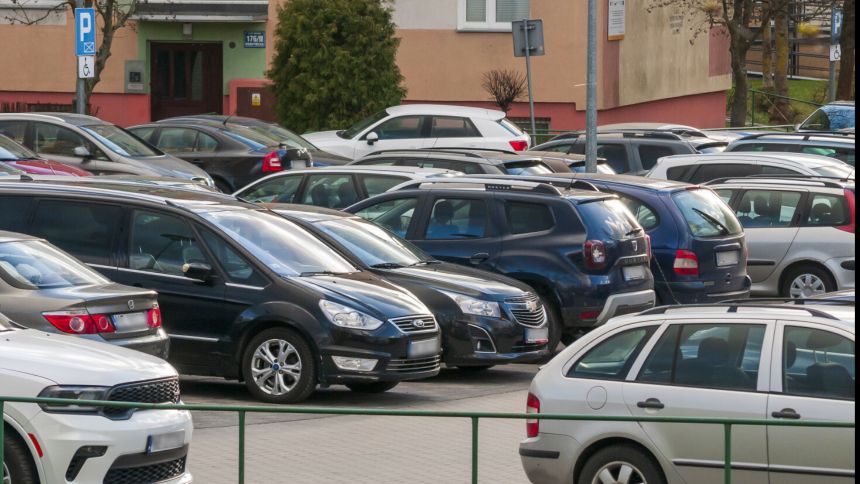 Parking pełen samochodów osobowych, z budynkiem w tle. Zielony znak parkowania w rogu.