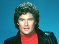 David Hasselhoff z kręconymi włosami w skórzanej kurtce i czerwonym swetrze na tle niebieskim.