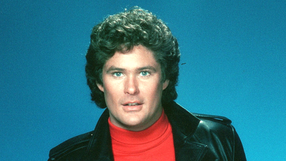 David Hasselhoff z kręconymi włosami w skórzanej kurtce i czerwonym swetrze na tle niebieskim.