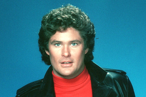 David Hasselhoff z kręconymi włosami w skórzanej kurtce i czerwonym swetrze na tle niebieskim.