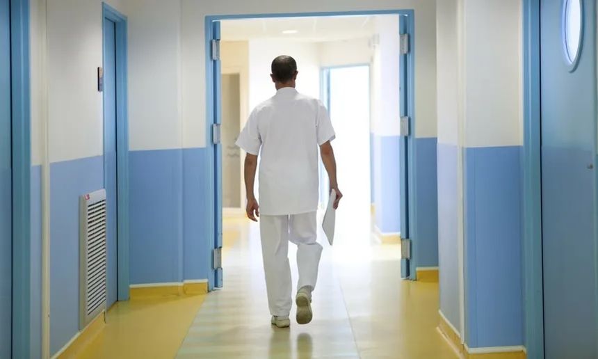Lekarz w białym stroju idzie korytarzem szpitalnym, z dokumentem w ręku. Jasne kolory ścian.