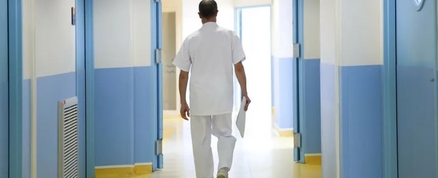 Lekarz w białym stroju idzie korytarzem szpitalnym, z dokumentem w ręku. Jasne kolory ścian.