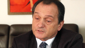 Mężczyzna w garniturze, z krótkimi włosami, mówi, siedząc na kanapie. W tle kolorowy obraz.
