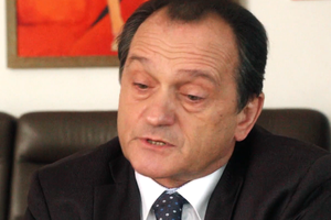 Mężczyzna w garniturze, z krótkimi włosami, mówi, siedząc na kanapie. W tle kolorowy obraz.