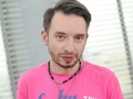 Mężczyzna w różowej koszulce z biżuterią, stojący w jasnym pomieszczeniu z oknami.