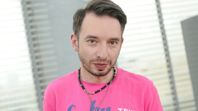 Mężczyzna w różowej koszulce z biżuterią, stojący w jasnym pomieszczeniu z oknami.