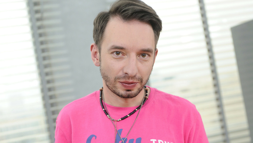Mężczyzna w różowej koszulce z biżuterią, stojący w jasnym pomieszczeniu z oknami.
