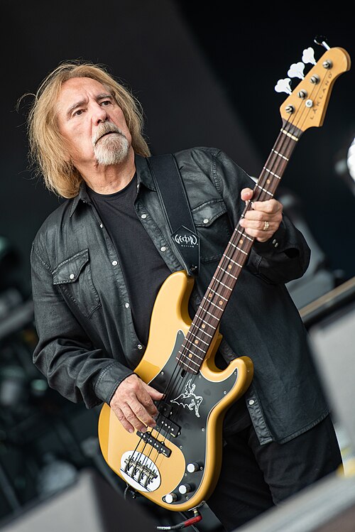 Geezer Butler. Mężczyzna w czarnej koszuli gra na basie, ma długie włosy i poważny wyraz twarzy.