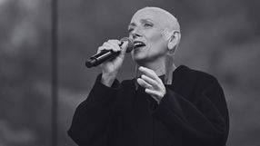 Kobieta z krótkimi, szarymi włosami śpiewa w mikrofon, ubrana w czarny płaszcz.