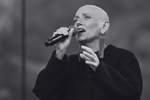 Kobieta z krótkimi, szarymi włosami śpiewa w mikrofon, ubrana w czarny płaszcz.
