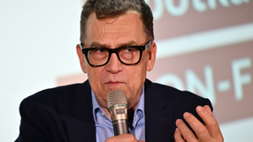 Mężczyzna w okularach, trzyma mikrofon, wygląda na zaangażowanego w rozmowę.
