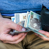 Ręka trzymająca banknoty w portfelu, na tle spodni w kolorze beżowym.