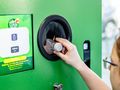 Osoba wrzucająca butelkę do zielonego recyklomatu. W tle wyświetlacz z informacjami.
