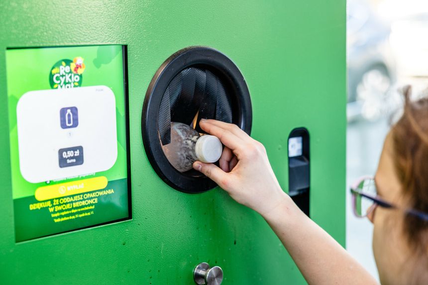 Osoba wrzucająca butelkę do zielonego recyklomatu. W tle wyświetlacz z informacjami.