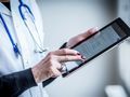 Osoba w białym fartuchu medycznym używa tabletu, przyglądając się ekranowi.