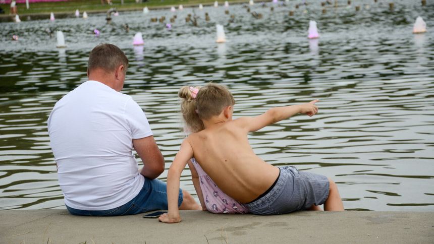 Mężczyzna, dziewczynka i chłopiec siedzą nad fontanną, chłopiec wskazuje w kierunku wody.