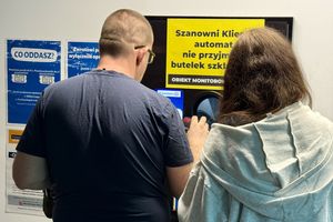 Dwoje ludzi przy automacie do zwrotu butelek, obok informacje o zasadach zwrotów.