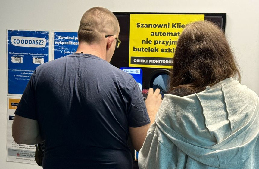 Dwoje ludzi przy automacie do zwrotu butelek, obok informacje o zasadach zwrotów.