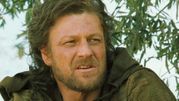 Na zdjęciu mężczyzna z długimi, nieco kręconymi włosami i brodą, ubrany w ciemną, zniszczoną odzież.