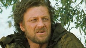 Na zdjęciu mężczyzna z długimi, nieco kręconymi włosami i brodą, ubrany w ciemną, zniszczoną odzież.