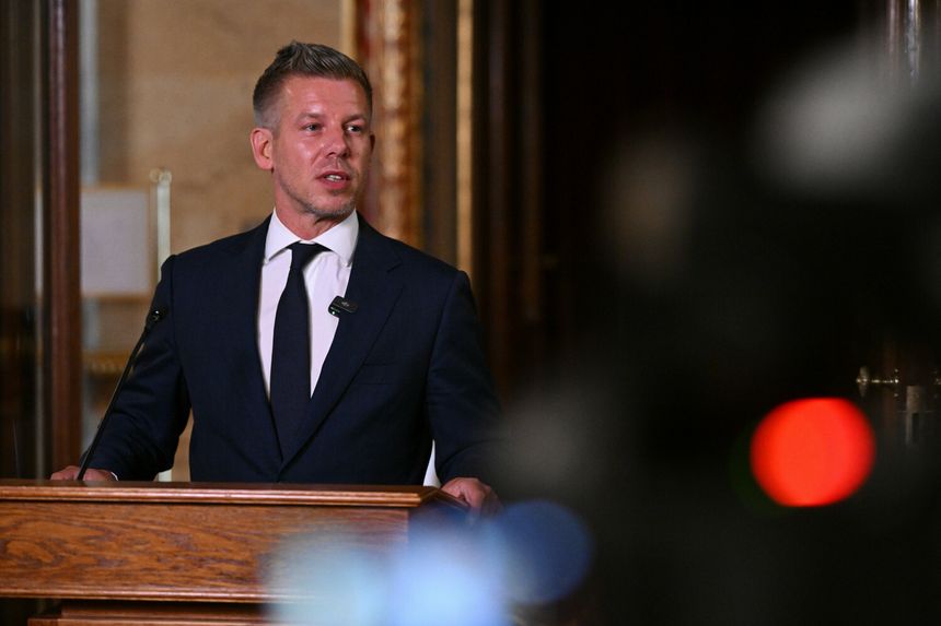 Mężczyzna w garniturze przemawia przy mównicy, w tle kamery i eleganckie wnętrze.