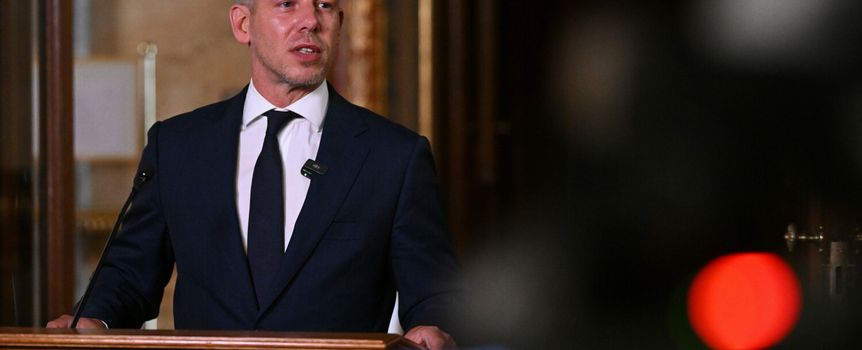 Mężczyzna w garniturze przemawia przy mównicy, w tle kamery i eleganckie wnętrze.