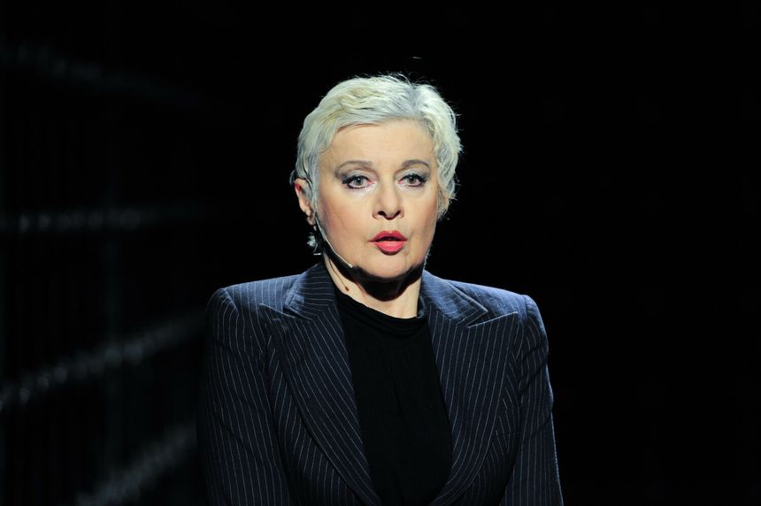 Kobieta z krótkimi, blond włosami w garniturze, na ciemnym tle, z mikrofonem.