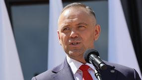 Mężczyzna w garniturze przemawia do mikrofonu, za nim widoczne flagi.