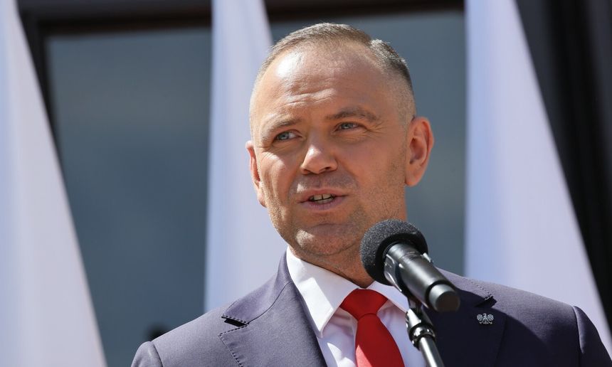 Mężczyzna w garniturze przemawia do mikrofonu, za nim widoczne flagi.