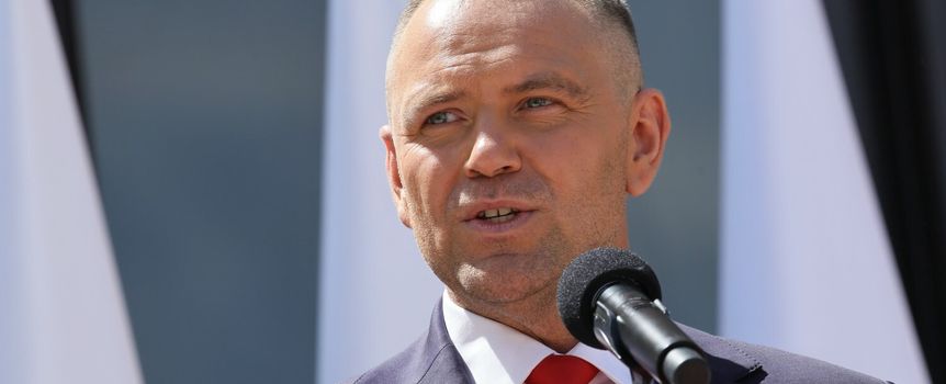 Mężczyzna w garniturze przemawia do mikrofonu, za nim widoczne flagi.