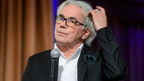 Robert Janowski. Mężczyzna z siwymi włosami w garniturze trzyma mikrofon, zastanawia się z gestem ręki.