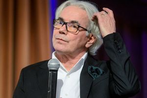 Robert Janowski. Mężczyzna z siwymi włosami w garniturze trzyma mikrofon, zastanawia się z gestem ręki.