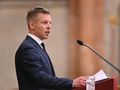 Mężczyzna w garniturze wygłasza przemówienie przy mównicy, obok szklanka z wodą.
