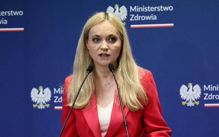 Kobieta w czerwonym żakiecie, mówiąca z mównicy, tło z logo Ministerstwa Zdrowia.