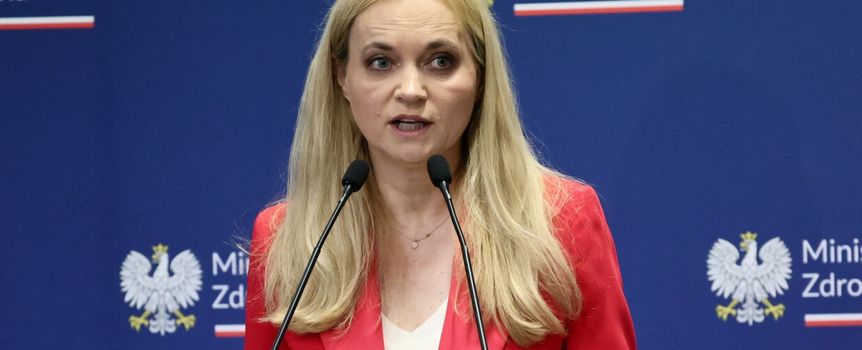 Kobieta w czerwonym żakiecie, mówiąca z mównicy, tło z logo Ministerstwa Zdrowia.