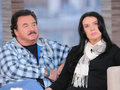 Dwoje ludzi na sofie w studiu telewizyjnym, mężczyzna w koszuli w kratę, kobieta w czerni.