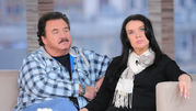 Dwoje ludzi na sofie w studiu telewizyjnym, mężczyzna w koszuli w kratę, kobieta w czerni.