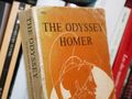 Okładka książki „The Odyssey” Homera na tle innych książek na półce.