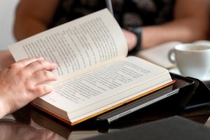 Osoba czyta książkę na stole, obok znajduje się filiżanka kawy i tablet.