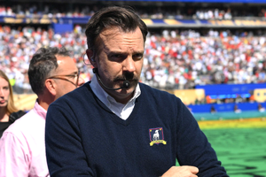 Mężczyzna w granatowym swetrze z mikrofonem, w tle tłum na stadionie.