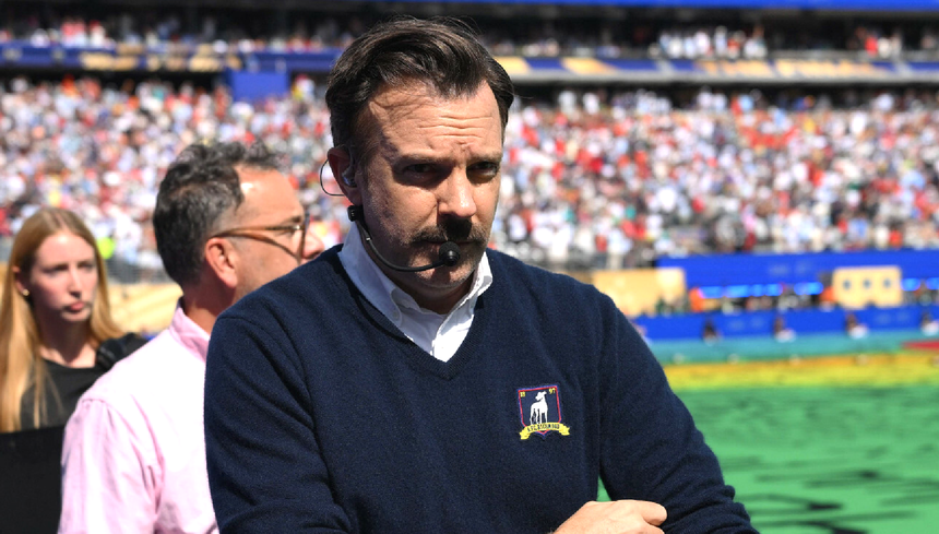 Mężczyzna w granatowym swetrze z mikrofonem, w tle tłum na stadionie.