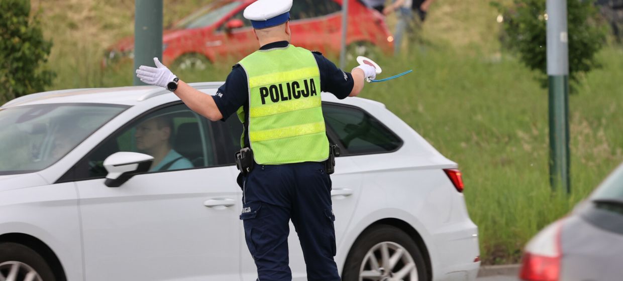 Policjant w odblaskowej kamizelce kieruje ruchem drogowym. W tle samochody i zieleń.