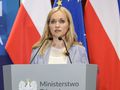Na pierwszym planie kobieta z blond włosami stoi przy mównicy z logo ministerstwa.