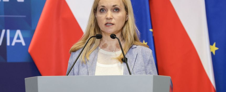 Na pierwszym planie kobieta z blond włosami stoi przy mównicy z logo ministerstwa.