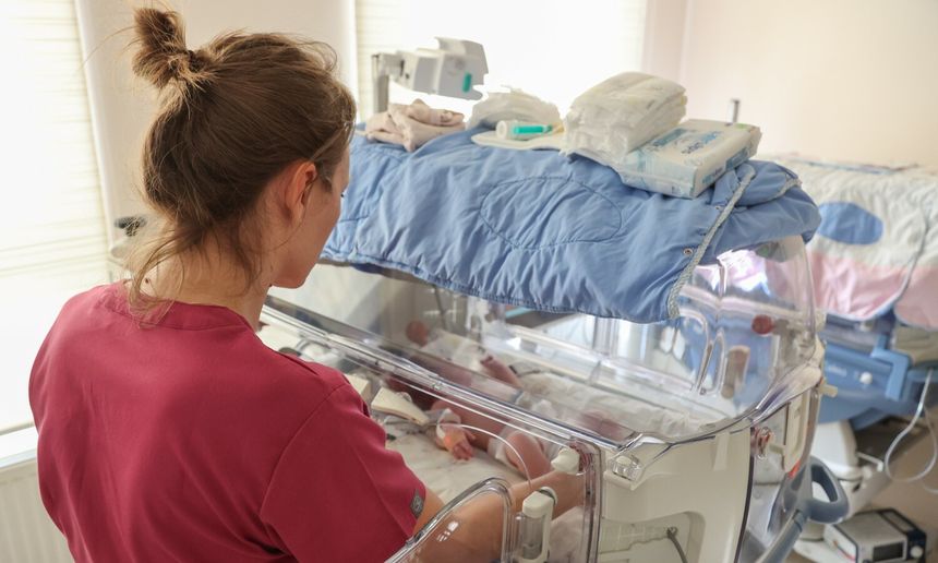 Zdjęcie przedstawia pielęgniarkę przy inkubatorze z noworodkiem w szpitalu.