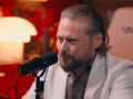 Krzysztof Stanowski z brodą w jasnym garniturze siedzi przy mikrofonie, rozmawiając w studiu.